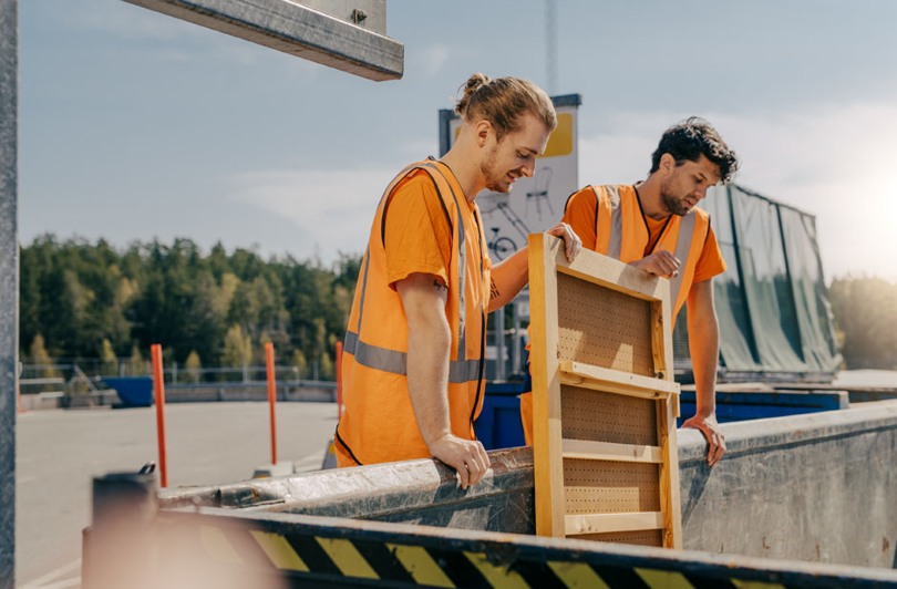 Två killar i varselkläder som slänger träavfall på en återvinningscentral