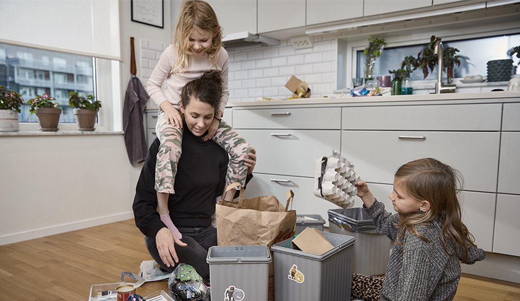 Kvinna som sitter på köksgolvet och sorterar sopor med sina barn. Ett barn sitter på golvet och ett barn sitter på kvinnans axlar.