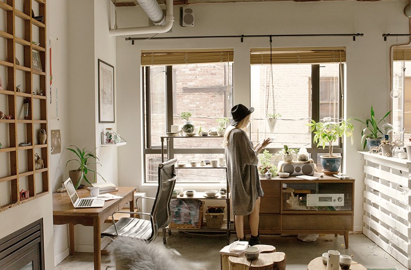 Person med hatt och klänning står i ett litet vardagsrum och tittar ut genom fönstret.